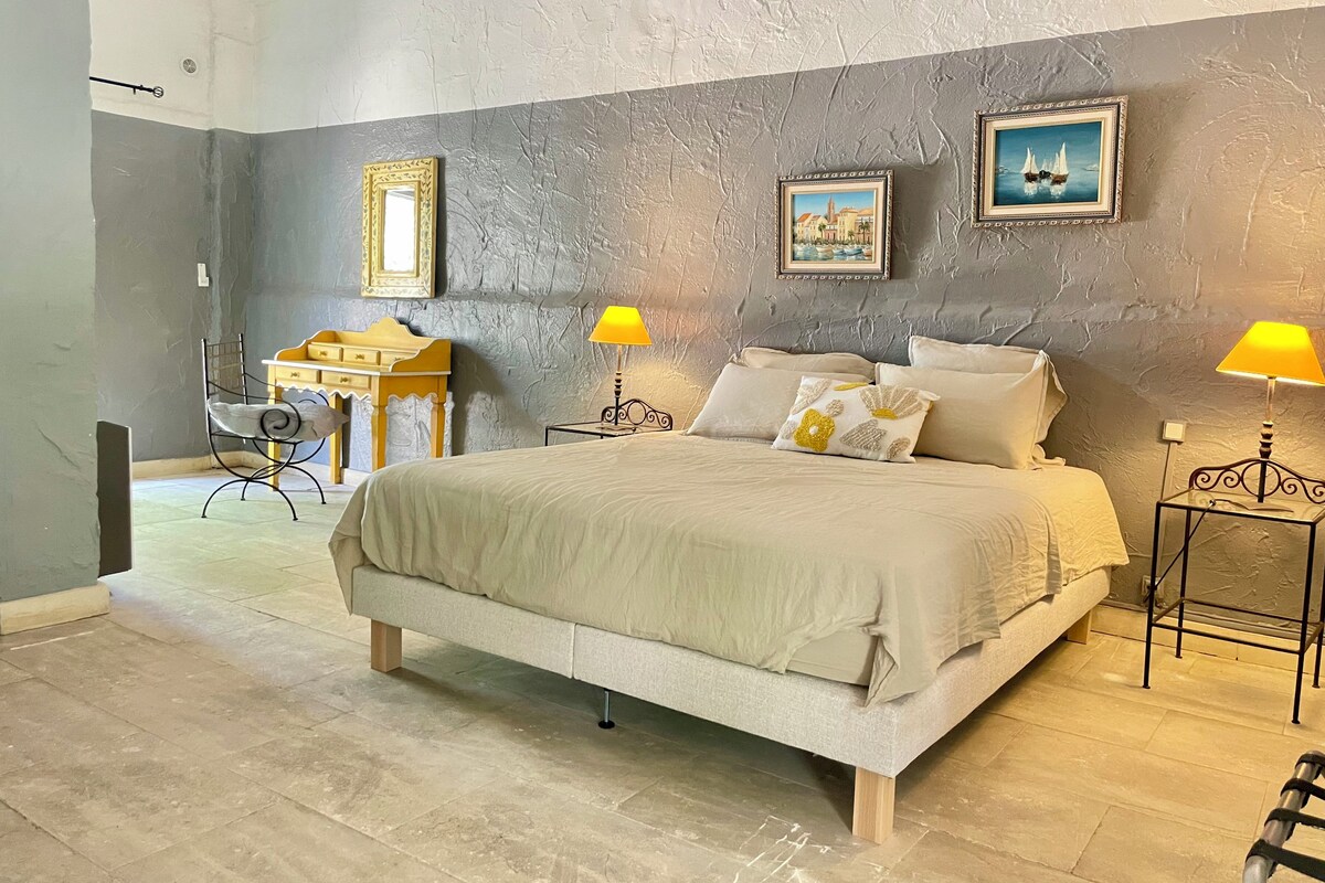 A bedroom with a large bed, two bedside tables with lamps, a chair, a small table, and two framed pictures on the wall.