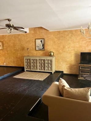 Quarto com piso de madeira escura, sofá bege, armário branco, ventilador de teto e televisão.