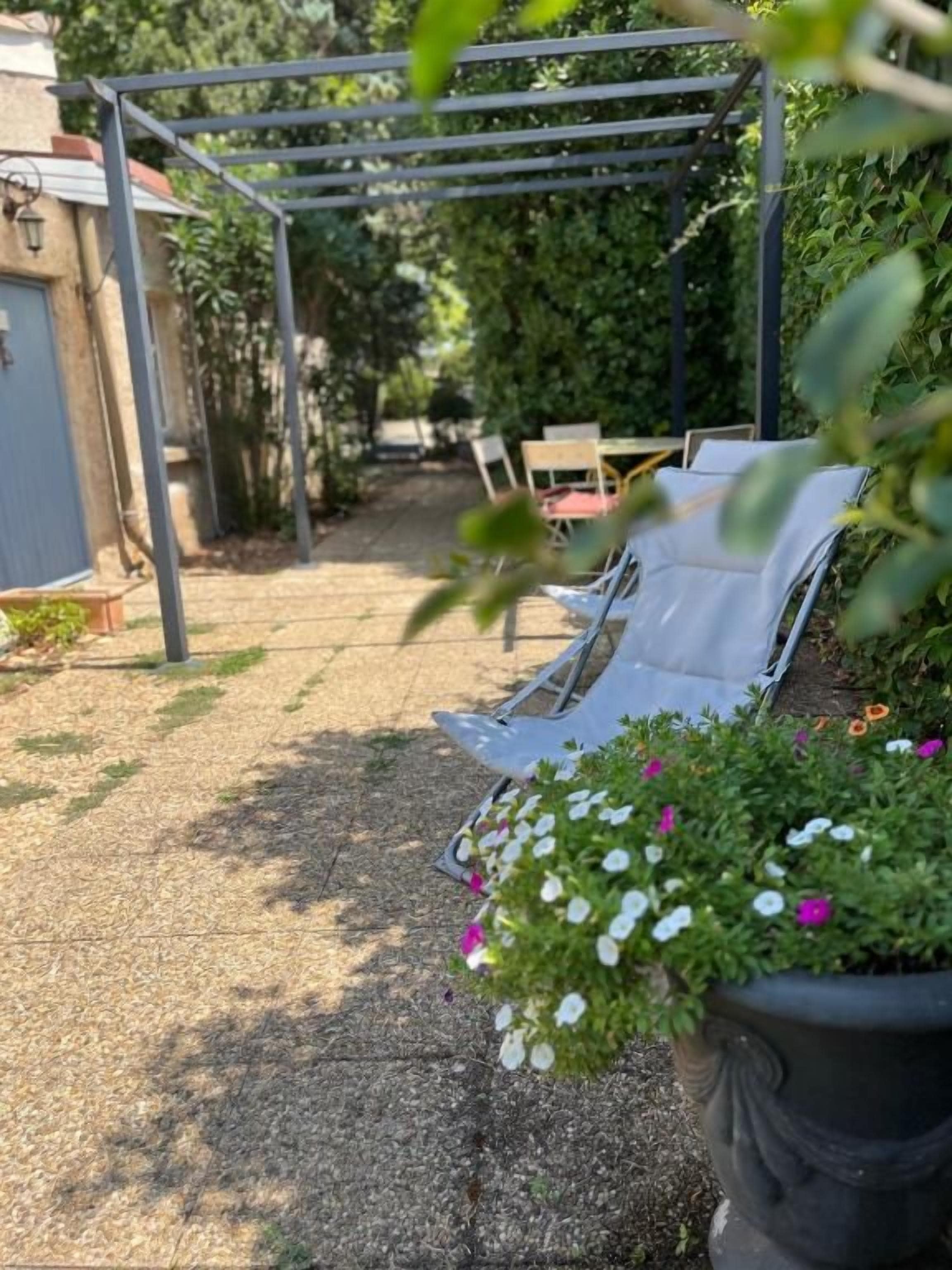 Un espace extérieur aménagé avec une pergola, des plantes en pot aux fleurs blanches et roses, et un chemin menant à un bâtiment.