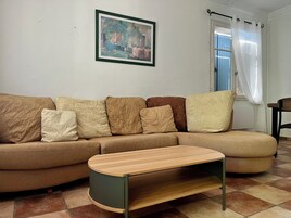 Una sala de estar con un sofá seccional, una mesa de centro de madera, un cuadro en la pared y una ventana con cortinas.