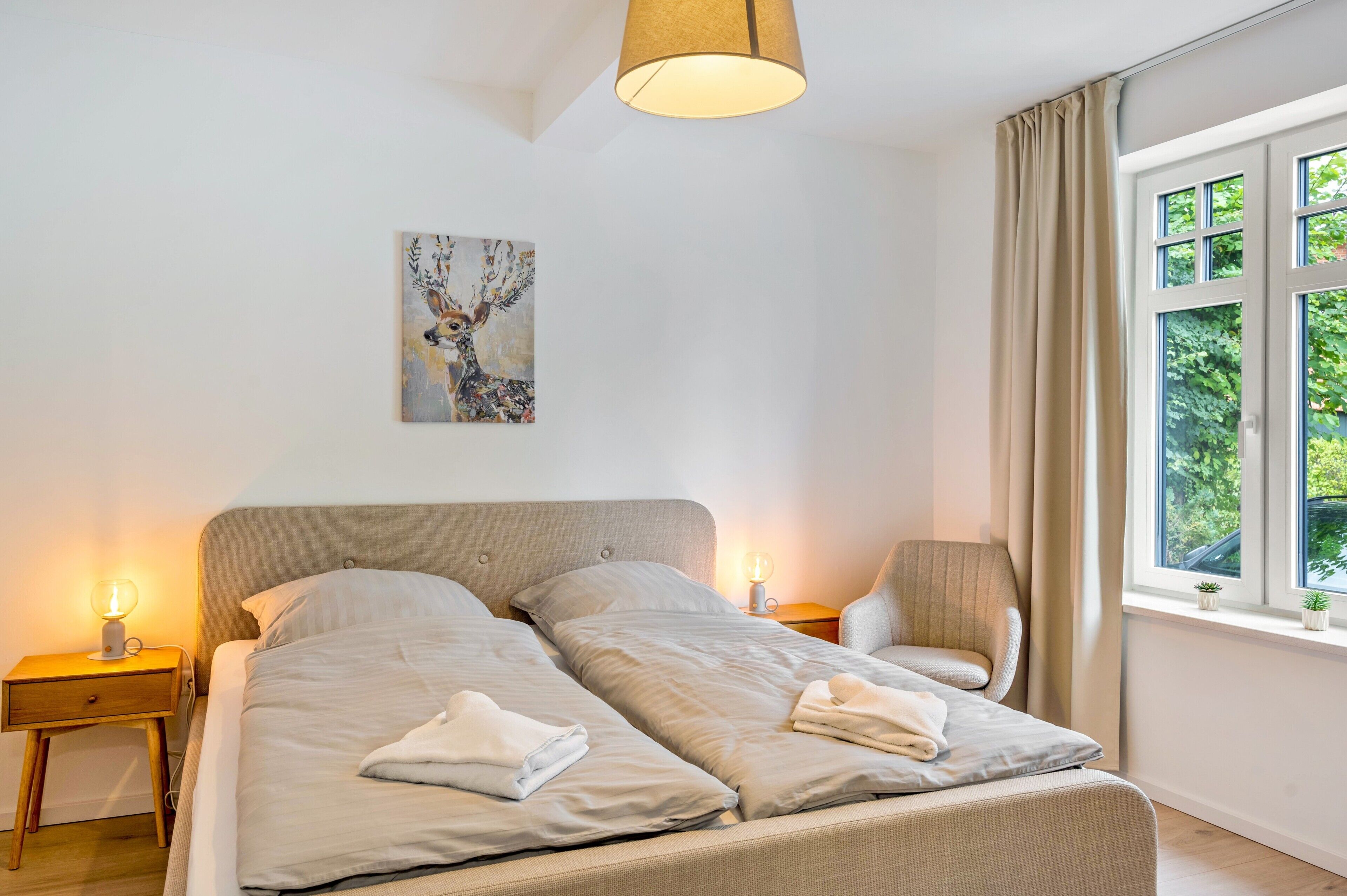 Une chambre à coucher avec un lit double, deux lampes de chevet, une chaise, une fenêtre avec des rideaux et un tableau au mur.