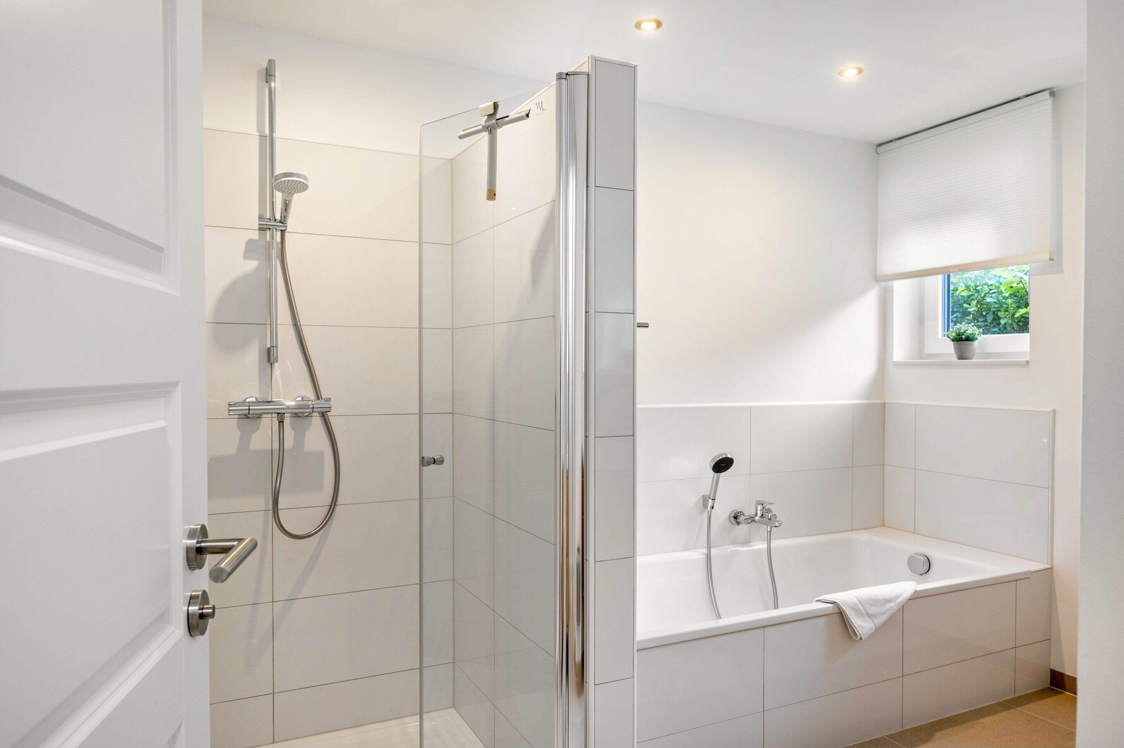 Ein modernes Badezimmer mit einer gläsernen Duschkabine, einer separaten Badewanne und einem Fenster mit Blick auf Grünflächen.