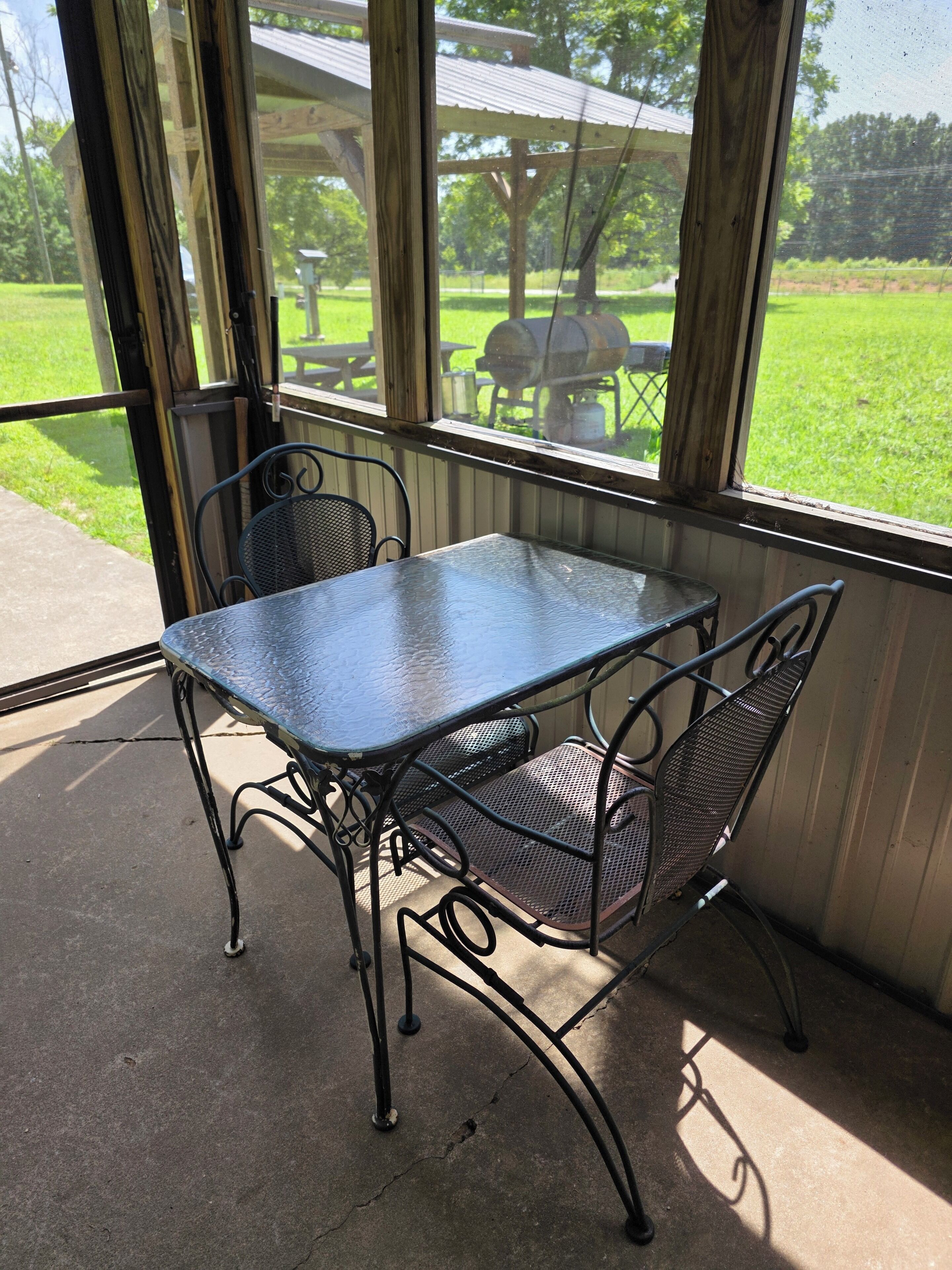 A covered patio area with a metal table and chairs, a view of a grassy park with barbecue equipment, and a wooden pergola structure.