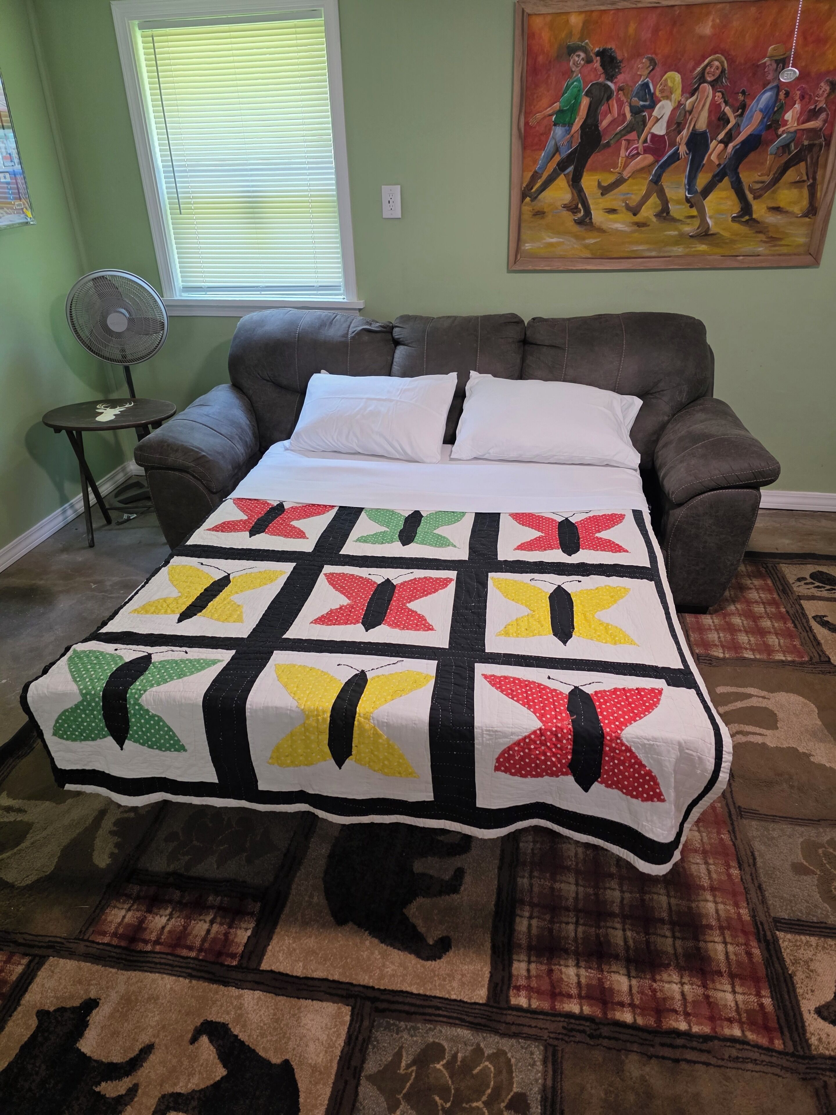 A bedroom with a large bed, a green wall, a painting, a fan, and a small table.