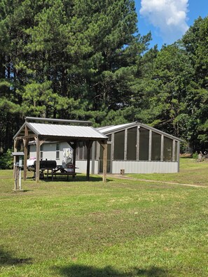 A small building with a metal roof and large windows, surrounded by a grassy field and dense trees. - Cabin with spacious yard north of Dover Ar.  National Forest, Moccasin Gap ATV (Dover)