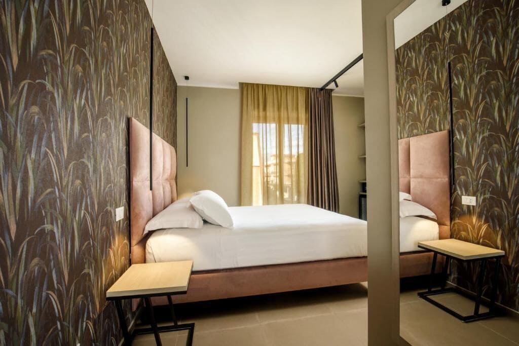 A hotel room with a bed, two bedside tables, a window with curtains, and a wall with a plant pattern.