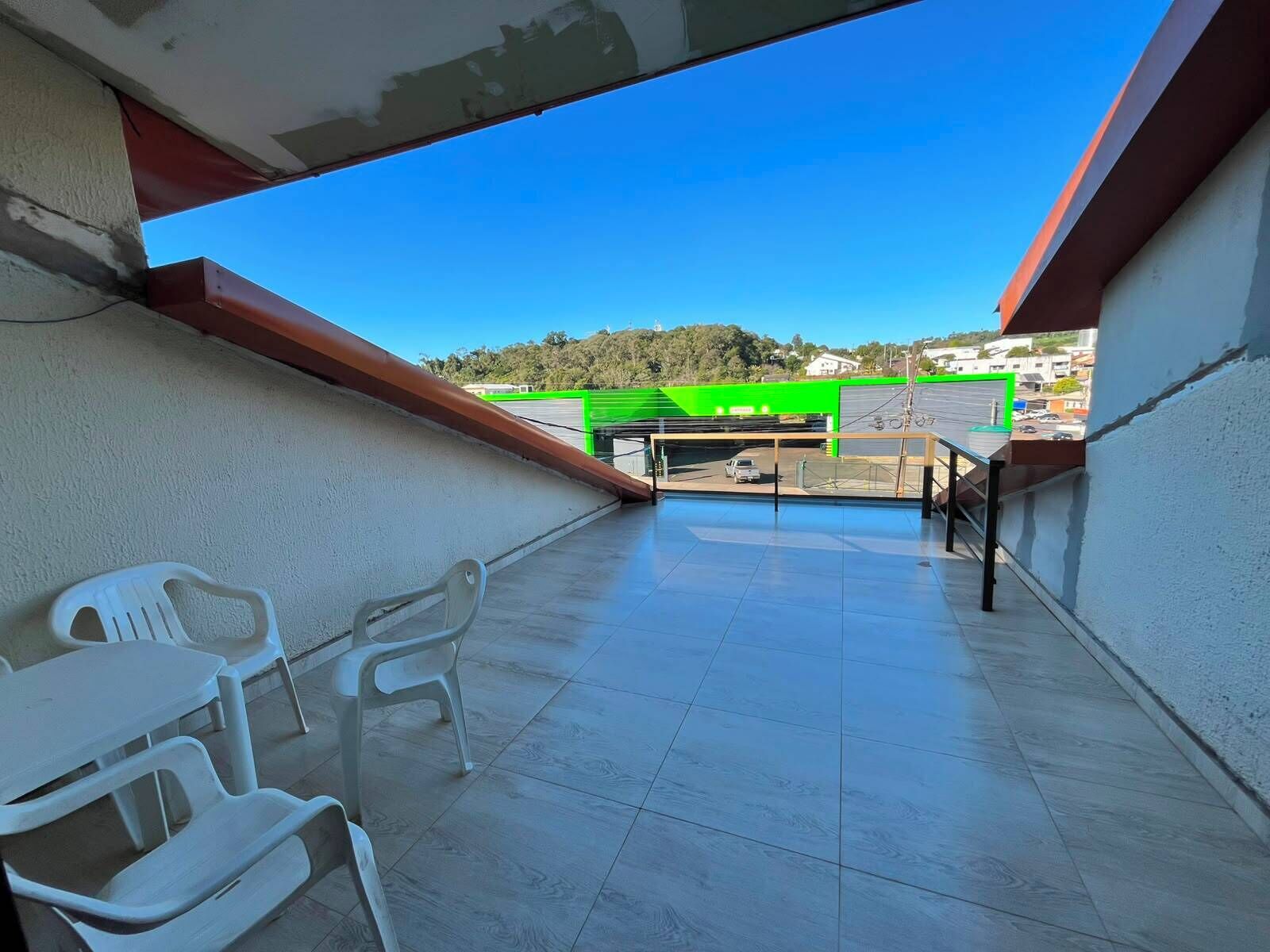 Uma varanda com cadeiras de plástico branco, chão de azulejos e vista para um estacionamento com estrutura verde.