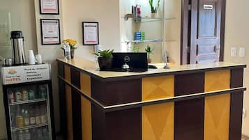 A hotel reception area with a reception desk, a refrigerator stocked with beverages, and a door labeled 'Estácio.'