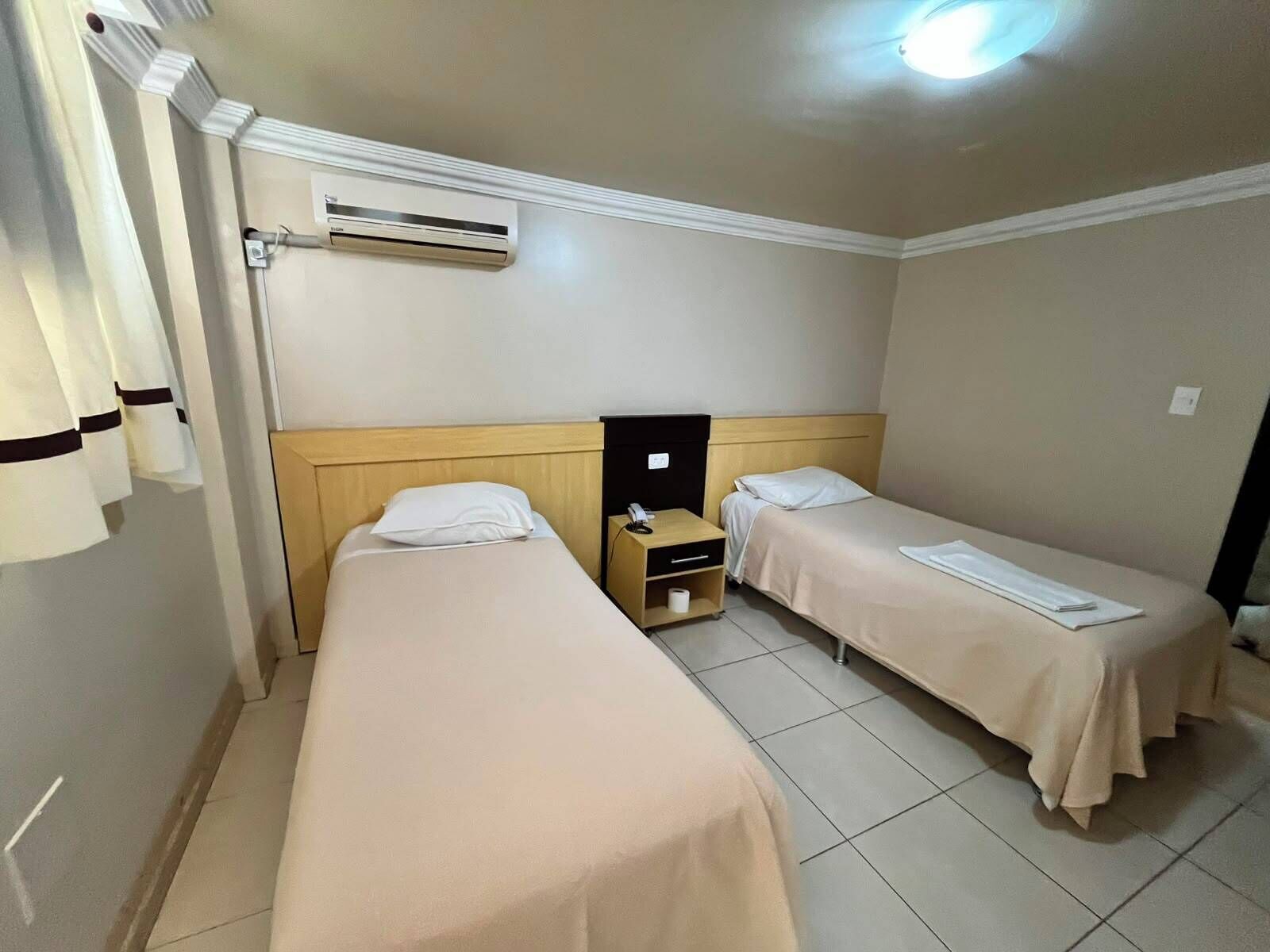 A hotel room with two beds, a nightstand, an air conditioning unit, and a ceiling light.