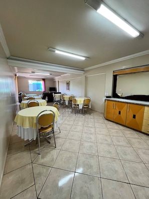 A dining area with tables and chairs, a kitchen area with cabinets, and a television screen in the background.