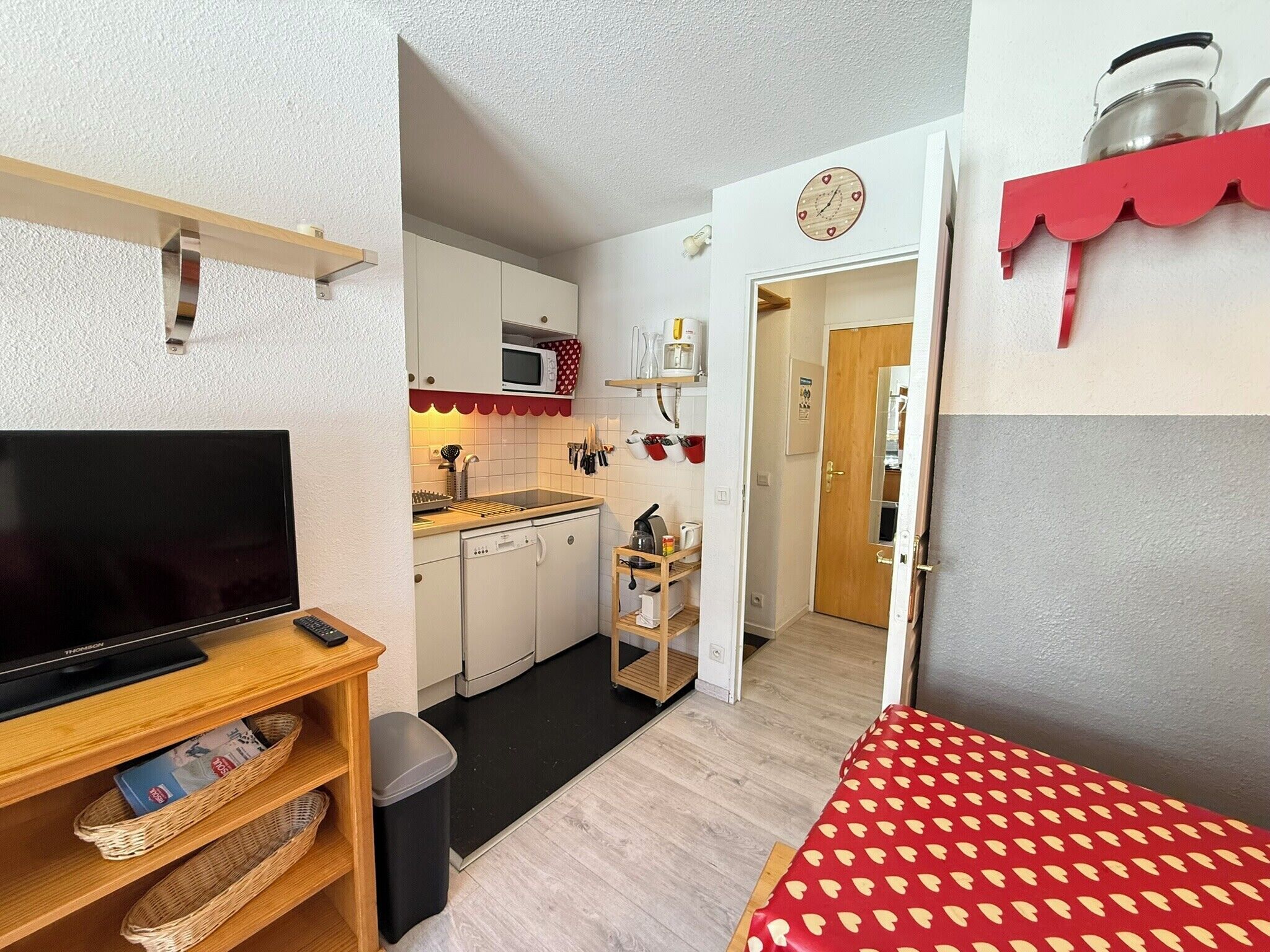 A compact living space with a television, kitchenette, and a red patterned area rug.