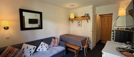 A compact living space with a sofa, a small table, a wall-mounted TV, and a kitchenette area.