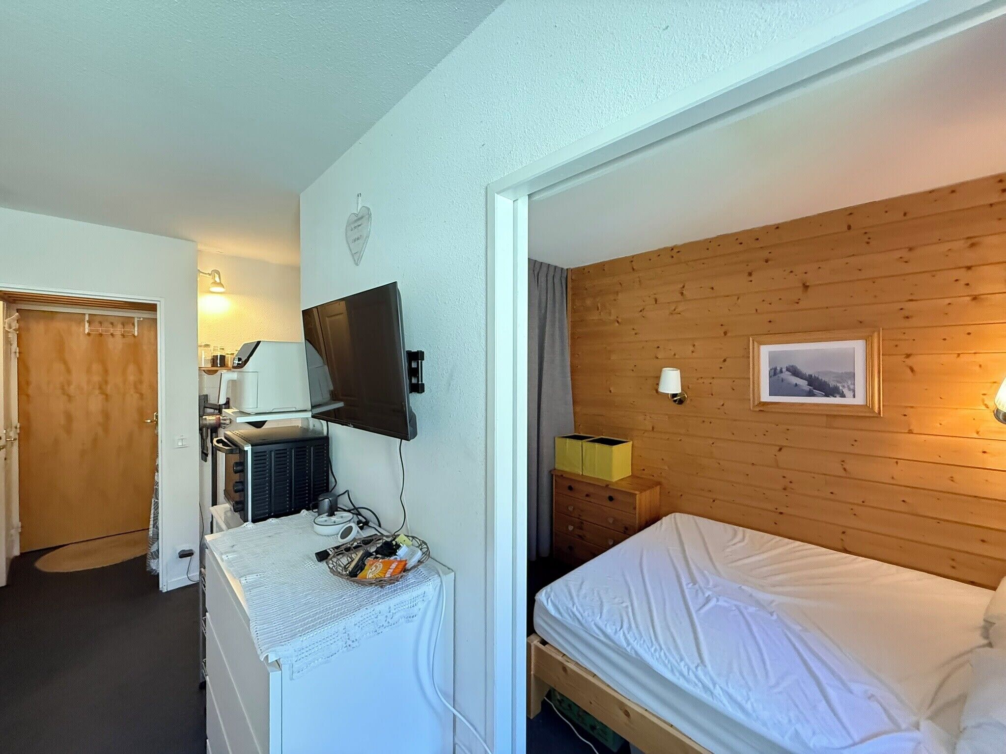 A room with a bed, a television mounted on the wall, a small bedside table, and a wooden wall panel.