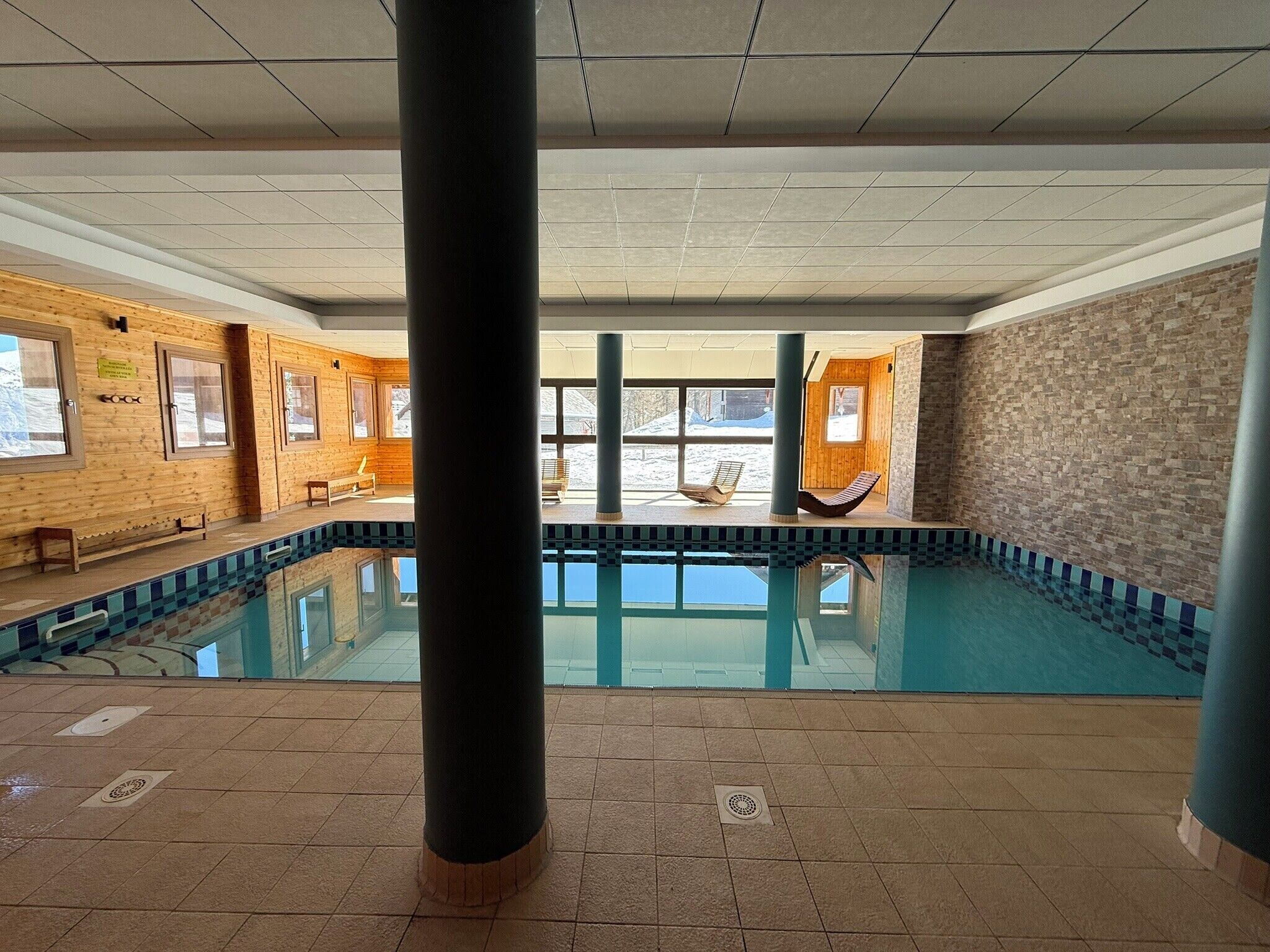 En indendørs swimmingpool med glasvægge, træbænke og flisegulv.