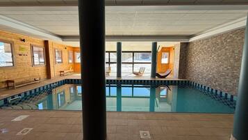 En indendørs swimmingpool med glasvægge, træbænke og flisegulv.