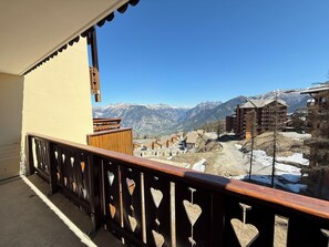 A balcony with a wooden railing, a view of snow-capped mountains, and buildings in the background. - Risoul • T2 6 pers • Piscine et sauna • Skis aux pieds (Risoul 1850)