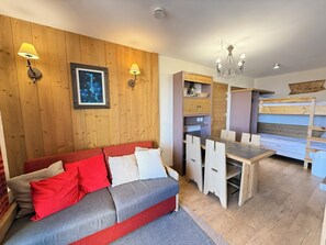 A cozy room with a wooden wall, a sofa with red and white cushions, a dining table with chairs, and a chandelier. - Risoul • T2 6 pers • Piscine et sauna • Skis aux pieds (Risoul 1850)
