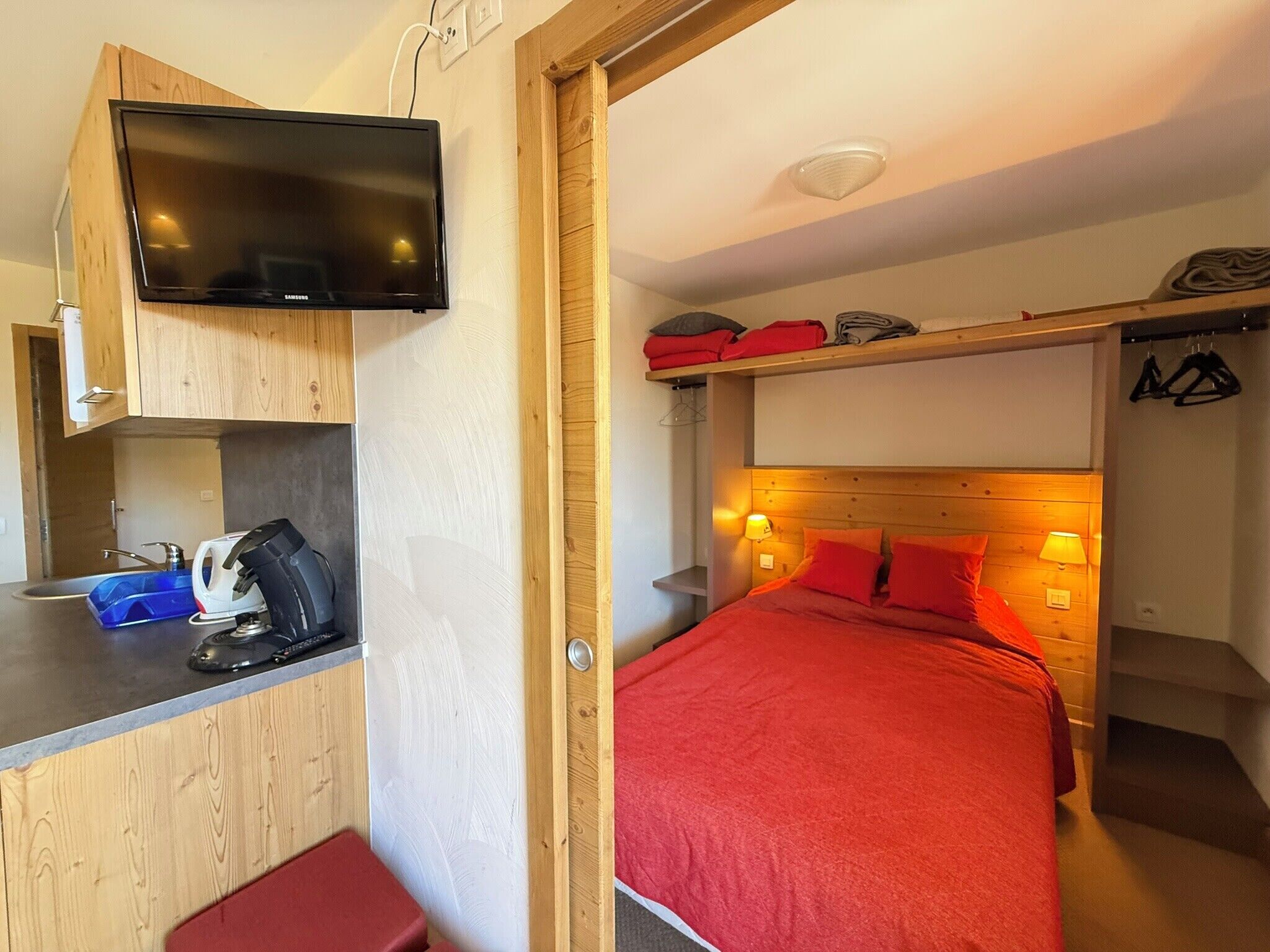 A compact room with a bed, a small kitchenette, and a television mounted on the wall.