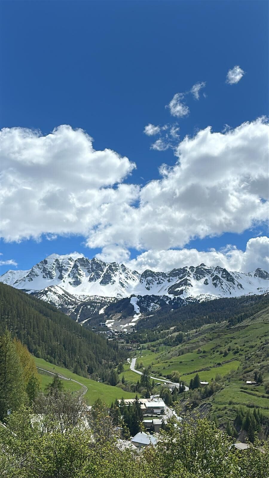 A mountainous landscape with a valley, a small settlement, and snow-capped peaks under a clear sky.
