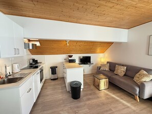 A modern kitchen with white cabinets, a wooden ceiling, a dining area with a grey sofa and patterned cushions, and a small side table. - Appart 6 pers - proche pistes et vue montagne (Vars)