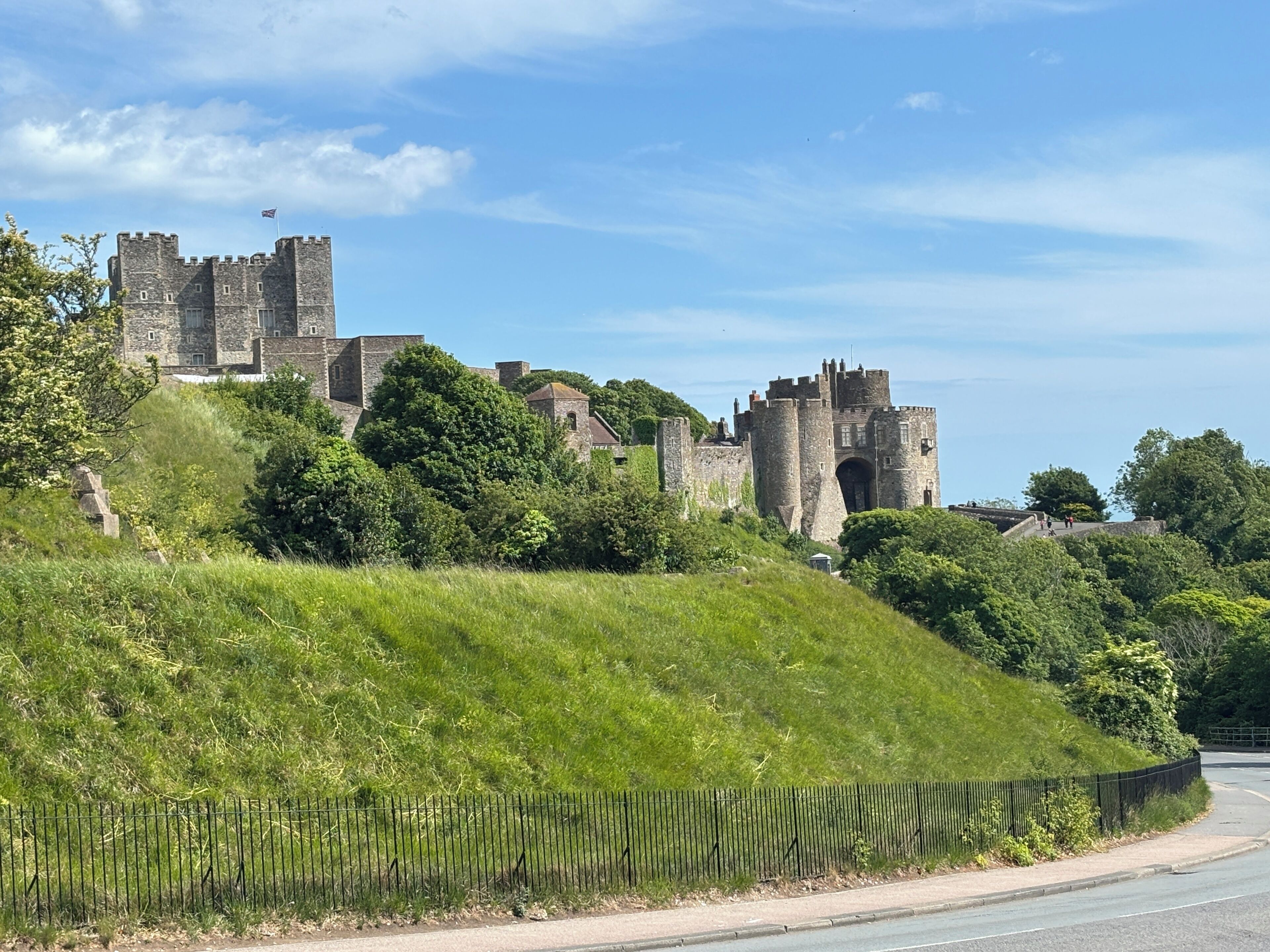 A historic stone castle with multiple towers and a flag, surrounded by lush greenery and a road with a fence in the foreground.