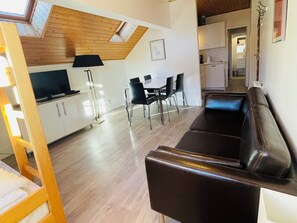 A modern living room with a wooden ceiling, a black leather sofa, a dining area with chairs, a TV on a cabinet, and a kitchen in the background. - Appart 4 pers - proche pistes et vue montagne (Vars)