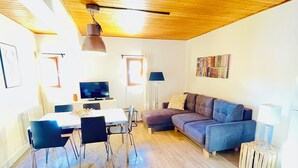 Uma sala de estar moderna com teto de madeira, um sofá cinza, uma mesa de jantar com cadeiras, uma televisão e uma estante de livros.