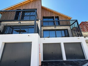 A multi-story building with a wooden facade, a balcony with black railings, and three garage doors.
