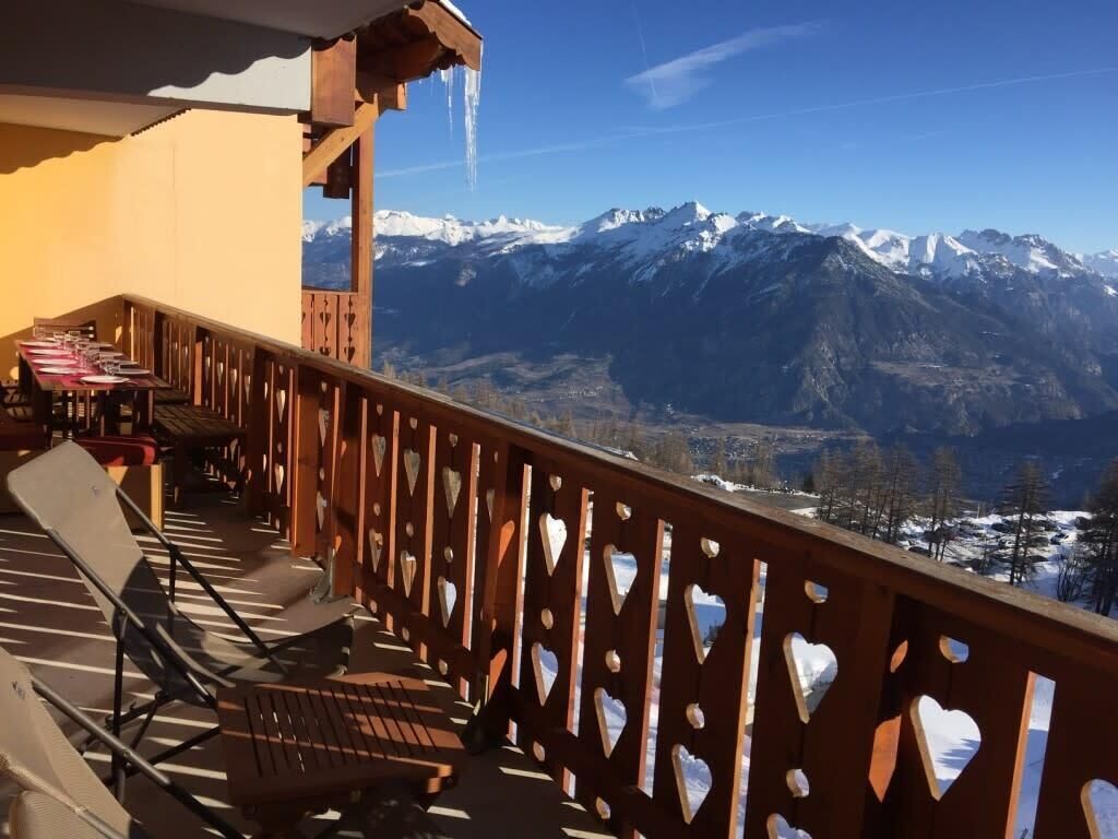 A balcony with a view of snow-capped mountains, wooden railings, and a table with chairs.