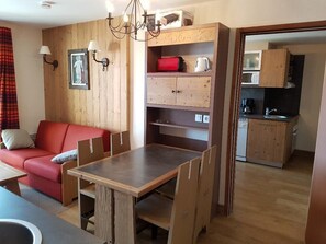 A room with a wooden wall, a red sofa, a wooden dining table with chairs, a kitchen area with cabinets, and a chandelier. - Appart 10 pers • Idéal Familles • Piscine & Sauna • Ski aux pieds (Risoul 1850)