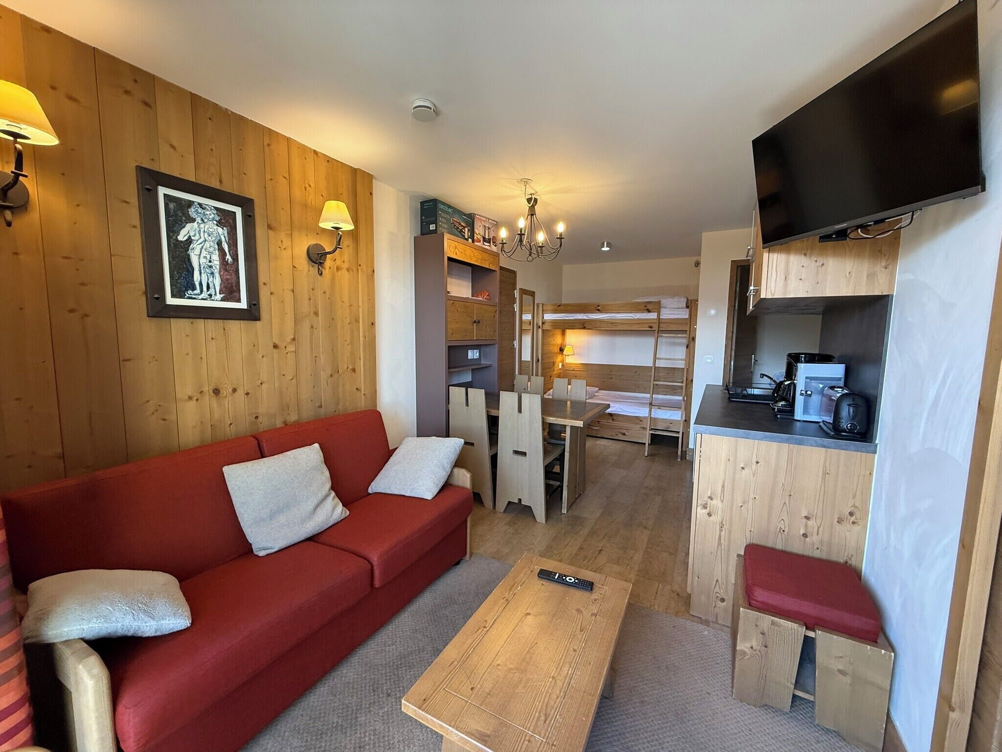A compact living space with a red sofa, a wooden coffee table, a kitchenette, and a television mounted on the wall.