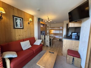 Ein gemütlicher, kleiner Wohnraum mit einem roten Sofa, einem Holztisch, einer Küchenzeile und einem an der Wand befestigten Fernseher.