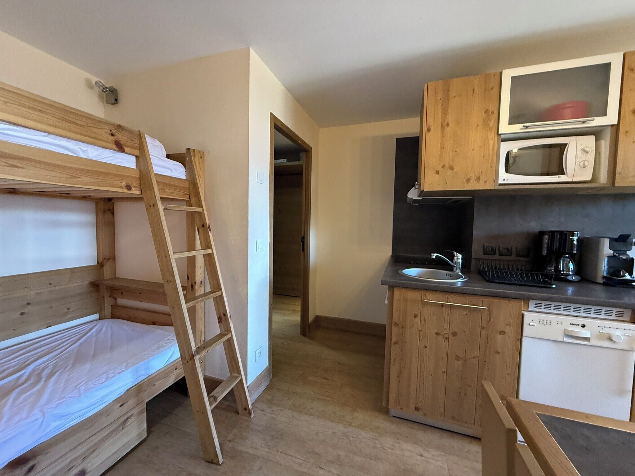 Habitación pequeña con literas, una escalera de madera, una kitchenette equipada con microondas, cafetera y lavavajillas.