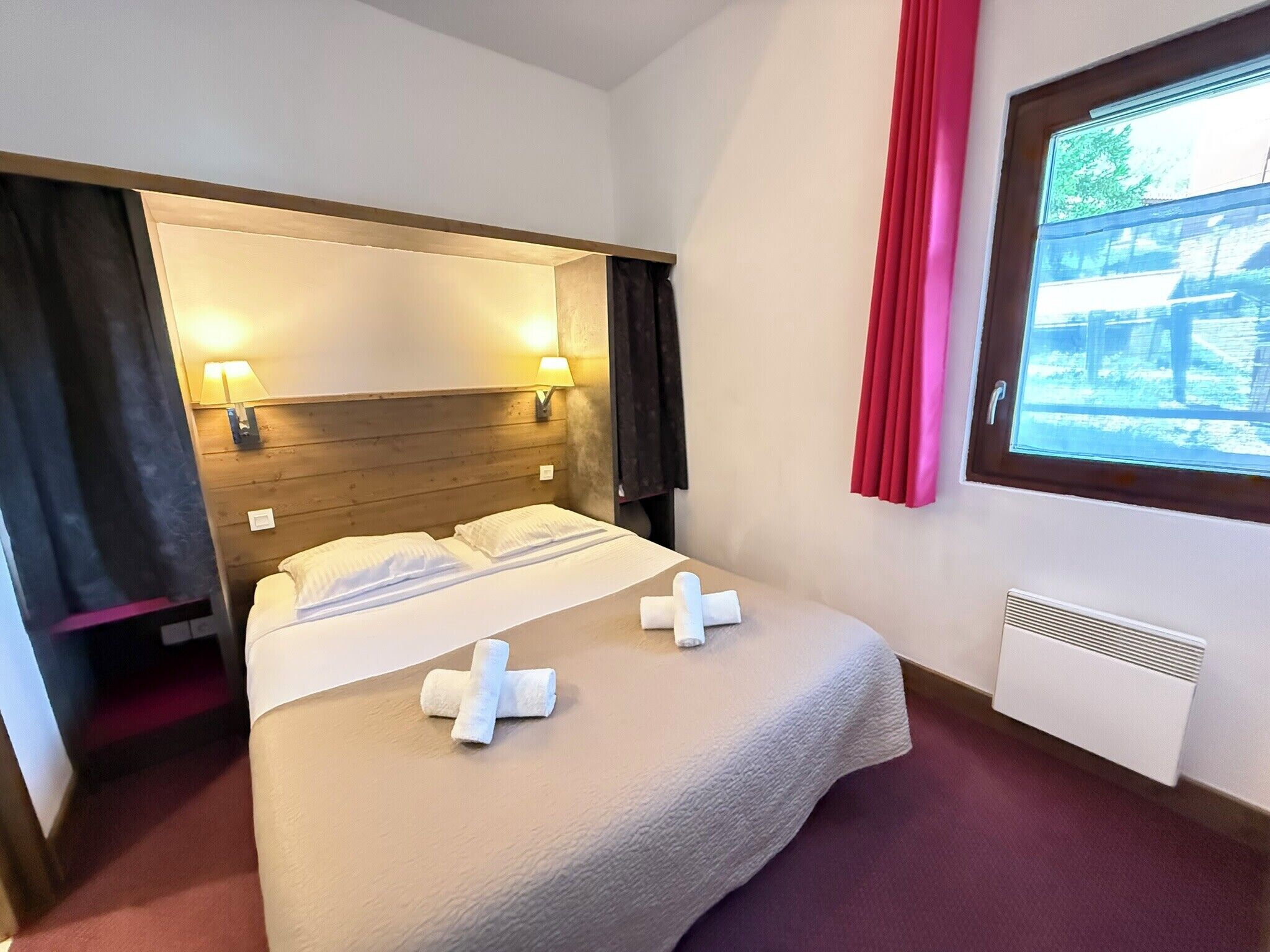 A hotel room with a bed, two bedside lamps, a window with curtains, and a radiator.