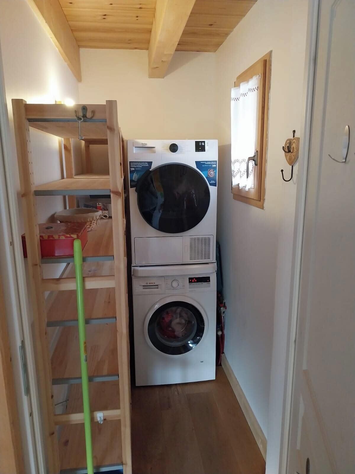 A laundry room with stacked washing machines, a wooden shelf unit, and a door with a coat hook.