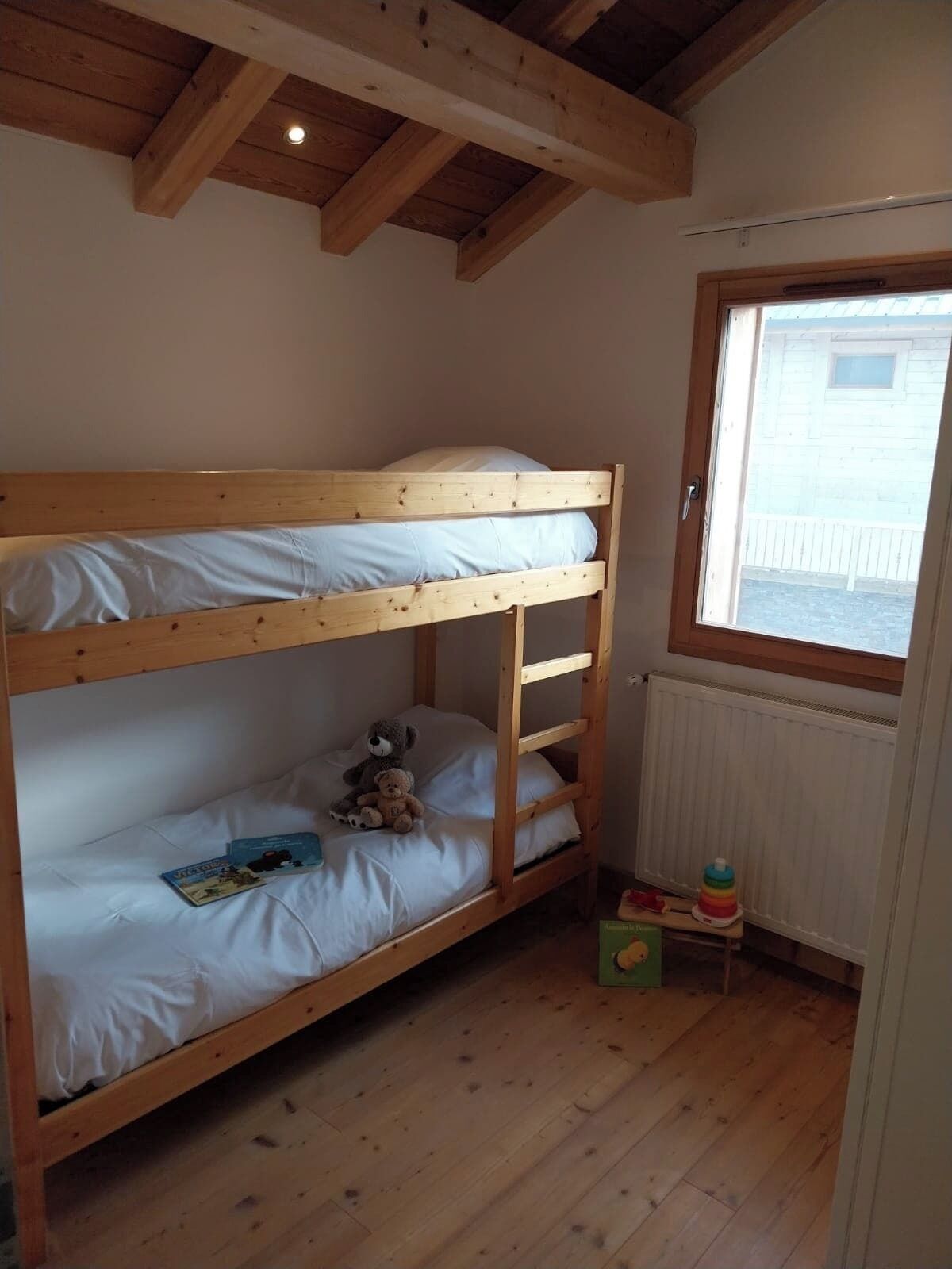 A bunk bed with a window, wooden floor, and a teddy bear on the lower bunk.