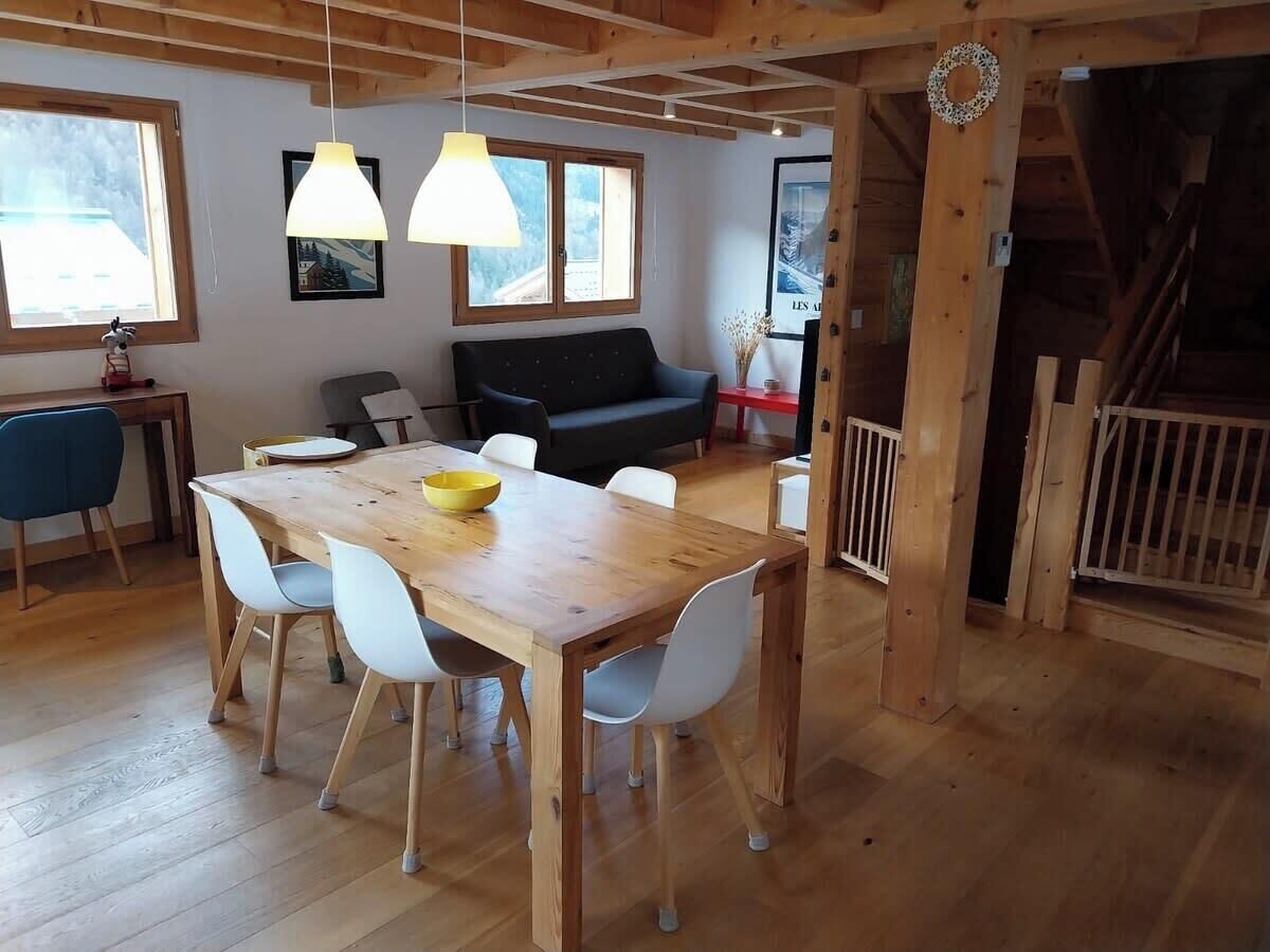 Une table à manger en bois avec des chaises, un canapé et des luminaires suspendus dans une pièce dotée de poutres apparentes et d’un escalier.