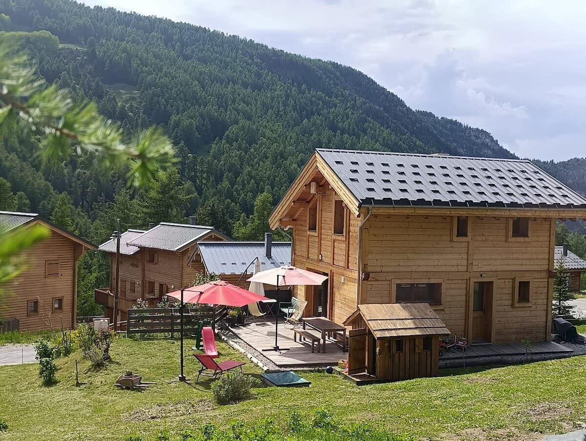 A wooden cabin with a patio area, surrounded by a forested mountain backdrop.