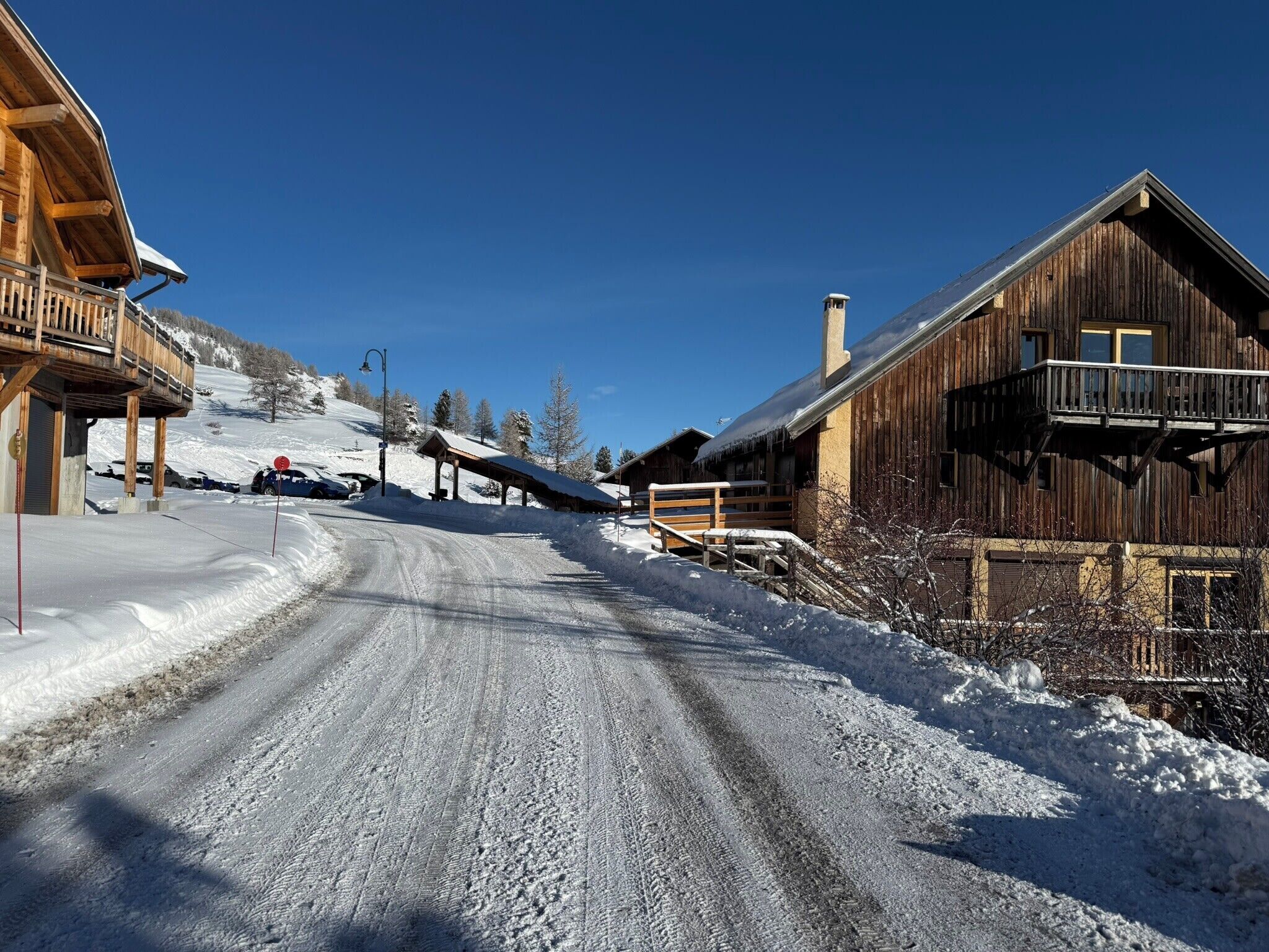Eine schneebedeckte Straße führt zu Holzbauten mit Balkonen, ein klarer blauer Himmel und ein Skiplatz im Hintergrund.