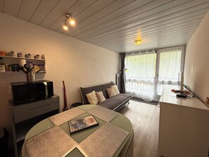 A modern room with a bed, a desk with a computer, a microwave, and a shelf with kitchen items.