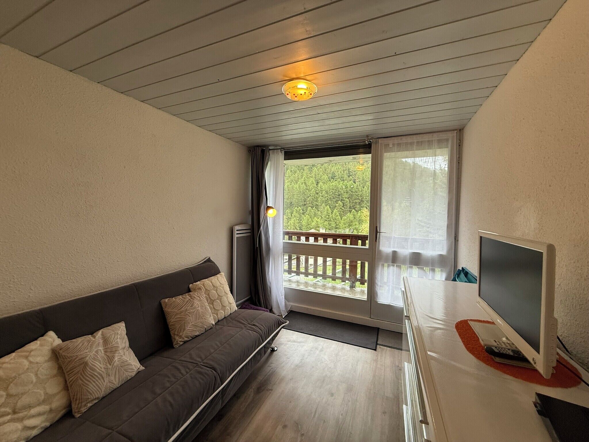Une pièce comprenant un canapé, un téléviseur, un balcon avec une rambarde et une porte donnant sur l’extérieur.