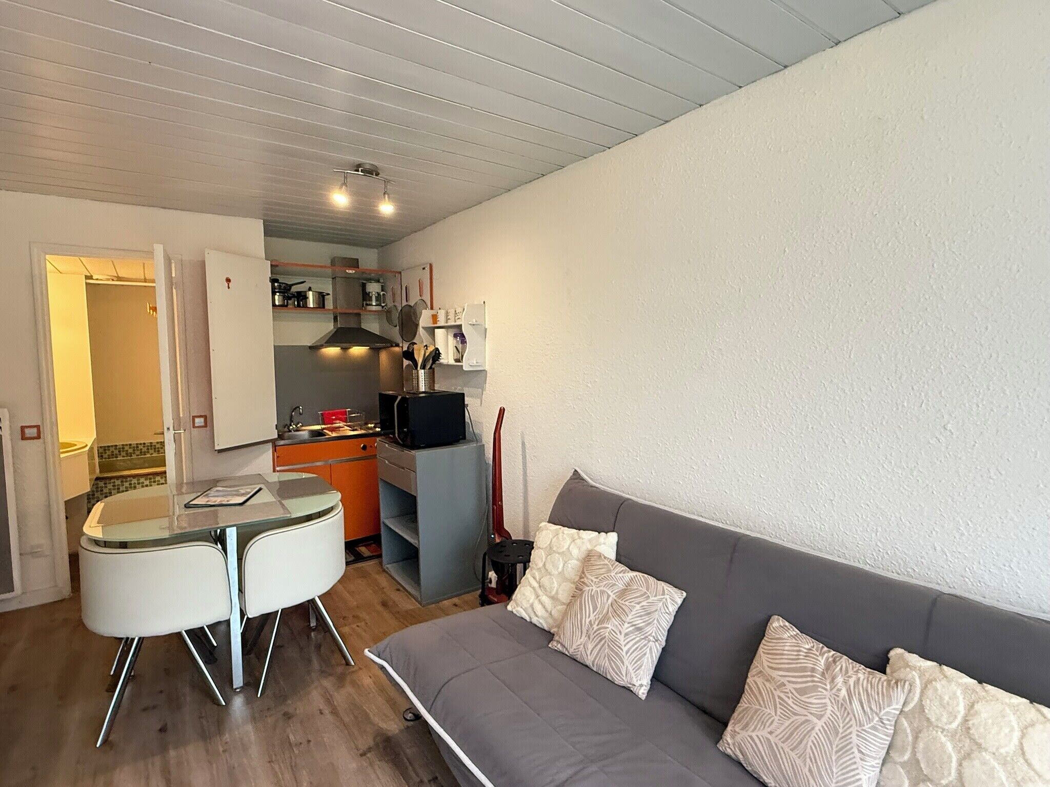 Een compact woongedeelte met een kitchenette, een eethoek met een glazen tafel en stoelen, en een grijze bank met gekleurde kussens.