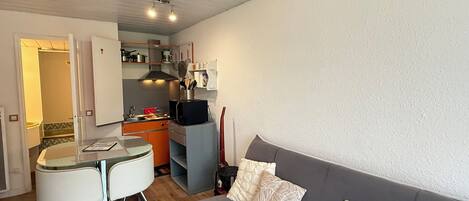 Een compact woongedeelte met een kitchenette, een eethoek met een glazen tafel en stoelen, en een grijze bank met gekleurde kussens.