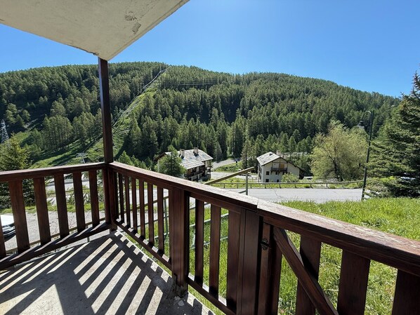 A wooden balcony with a view of a forested mountain and a small building with a sloped roof.