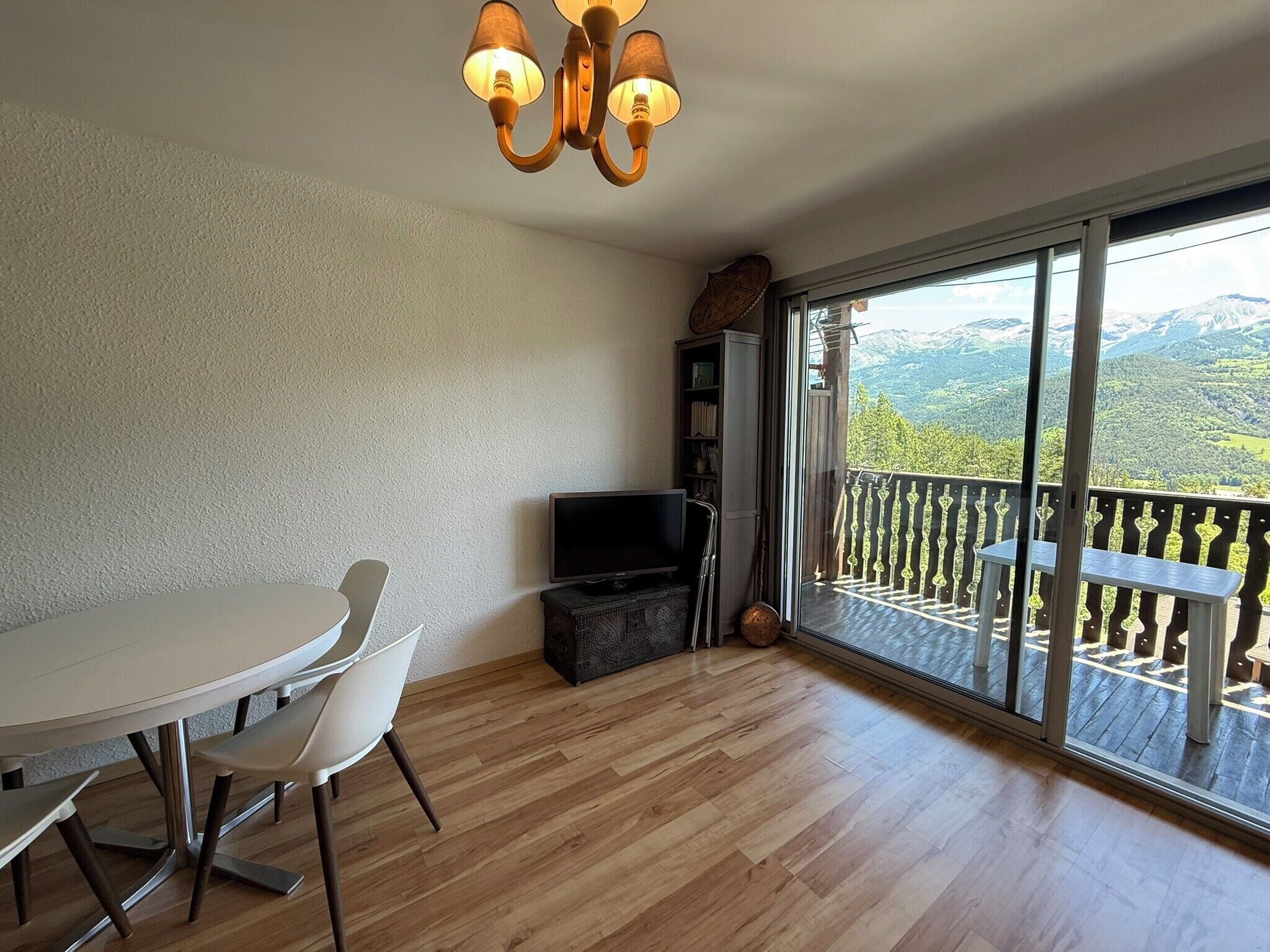 A room with a dining table, chairs, a television, and a large window with a balcony.