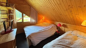 Ein gemütliches Schlafzimmer unter dem Dach mit schrägen Wänden, zwei Betten, einem Schreibtisch mit Lampe und einem Fenster mit Blick ins Freie.