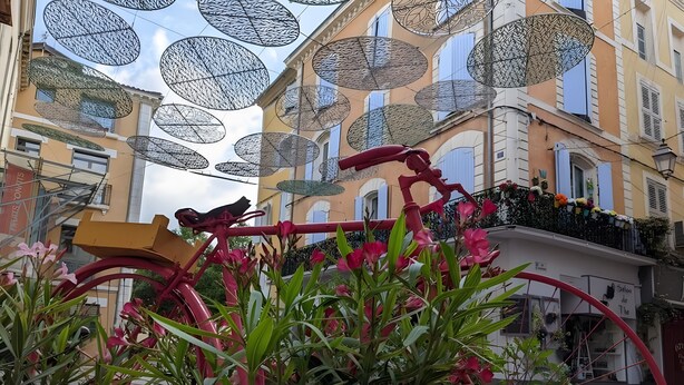 Una bicicleta rosa con estructura roja, rodeada de plantas verdes y flores rosadas.