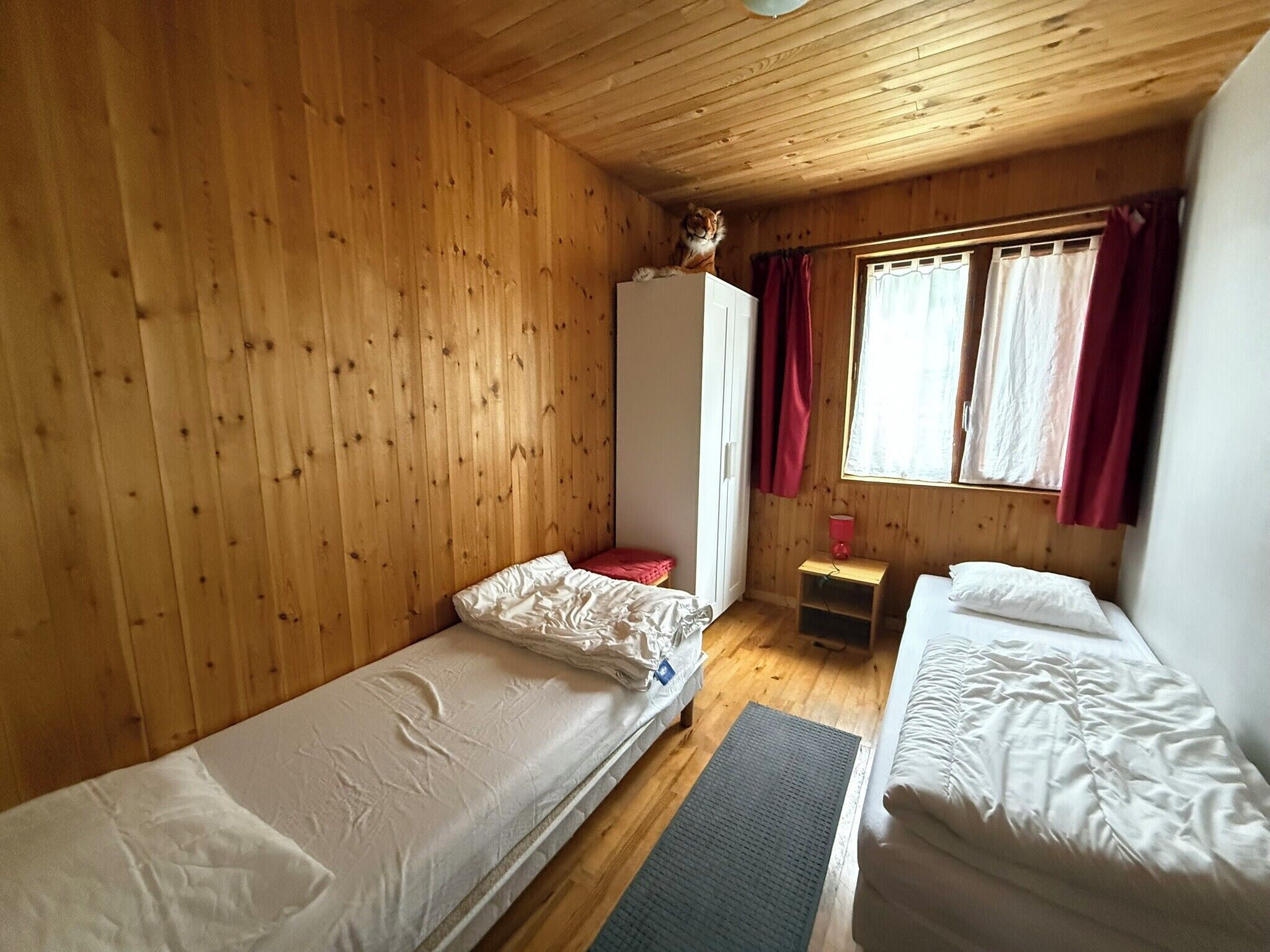 Ein Zimmer einer Holzhütte mit zwei Betten, einem Kleiderschrank und einem Fenster mit Vorhängen.