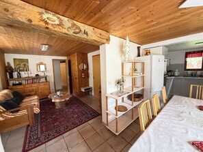 A cozy living room with a wooden ceiling, a dining area with a white table, and a kitchen with a refrigerator.