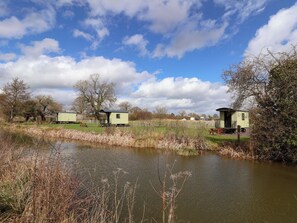A tranquil riverside setting with a row of small, light-colored cabins, a wooden bench, and a grassy area. - Quackin' Up (Whitchurch)